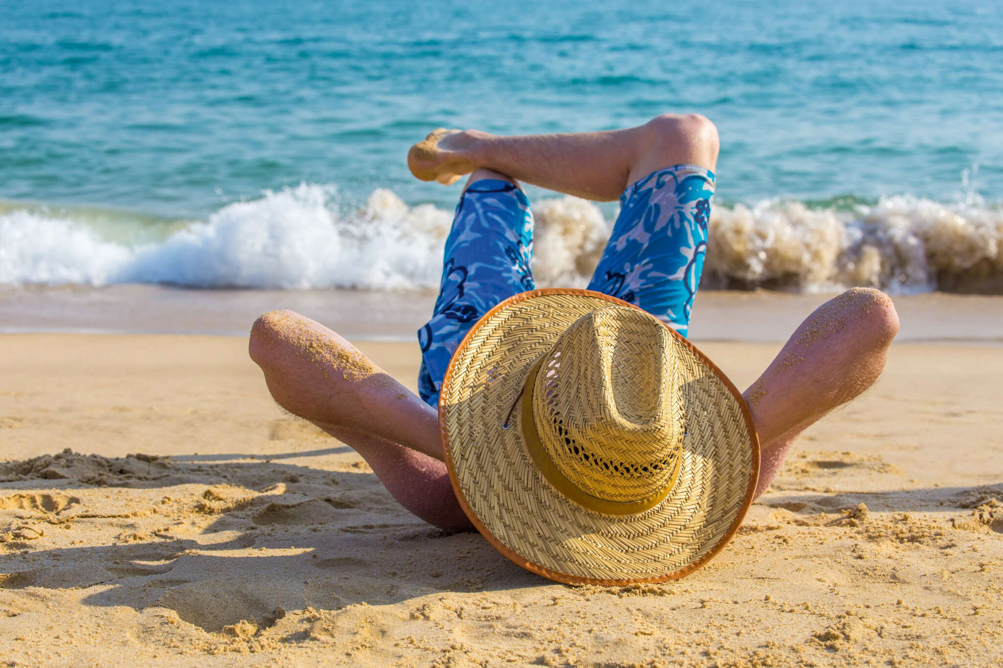 Ein Mann liegt am Strand mit übereinandergekreuten Beinen und hinter dem Kopf verschränkten Händen. Er trägt einen großen Strohhut, der sein Gesicht verdeckt