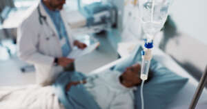 Patient liegt im Krankenbett und ist angeschlossen an Infusion, daneben steht ein Arzt und hat ein Papier in der Hand