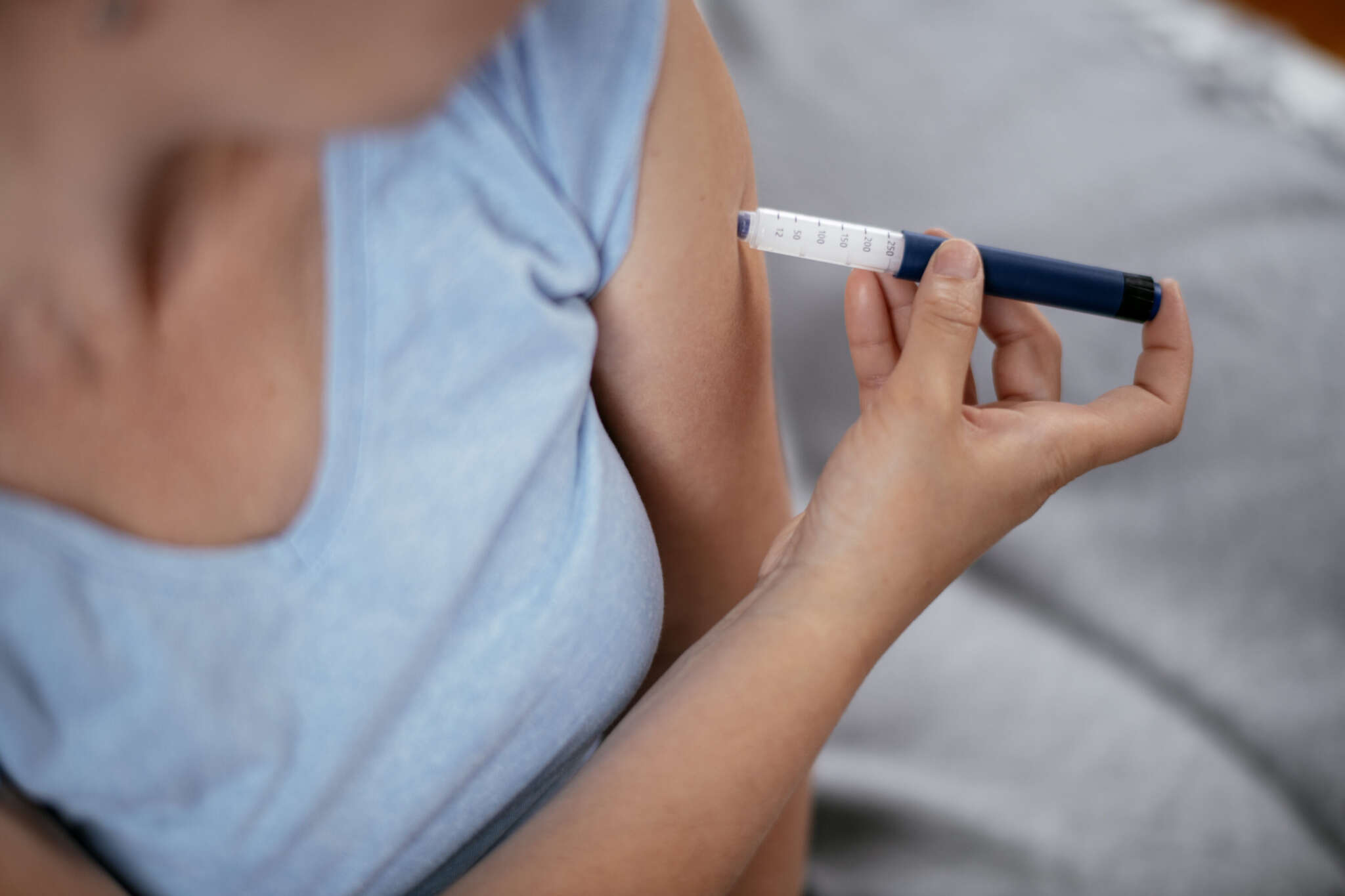 Frau in hellblauem T-Shirt spritzt sich in den linken Oberarm.