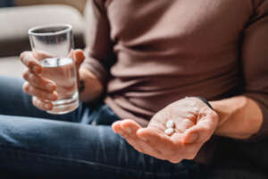 Person hält zwei weiße Tabletten in der linken Hand und in de rechten Hand ein Glas Wasser
