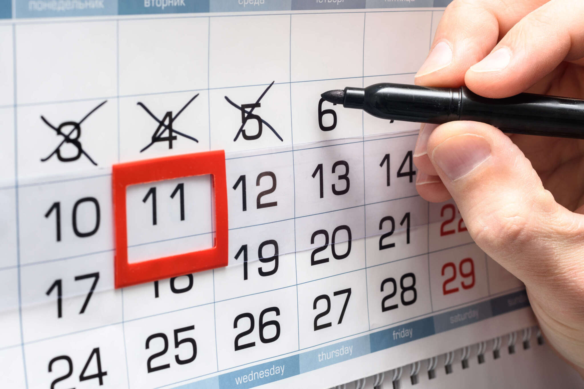 Eine Hand mit einem schwarzen Stift, ein Kalender mit einem roten Rahmen zur Markierung der Tage, einige Tage sind mit einem schwarzen Kreuz durchgestrichen