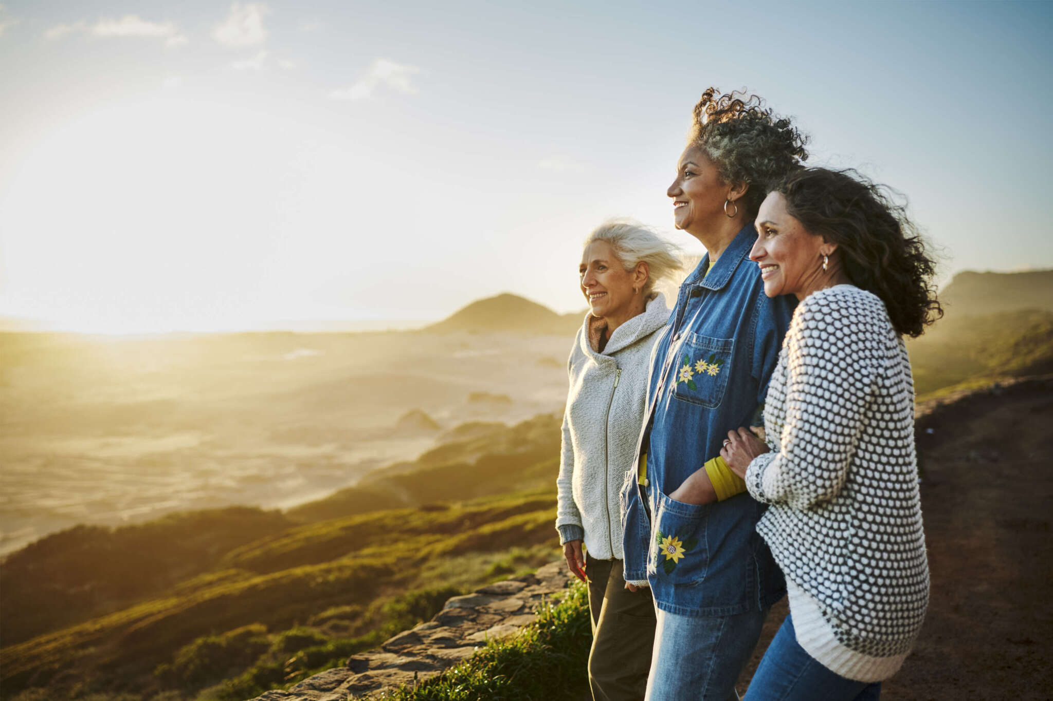 Drei Frauen stehen Arm in Arm und blicken auf eine Küstenlandschaft.