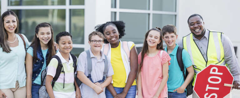 Schüler*innen stehen mit Lehrerin und Schulweghelfer vor einer Schule. Ein Schüler in der Mitte hat Down-Syndrom.